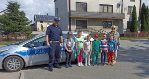 Policjant z dziećmi na tle radiowozu w tle budynki.