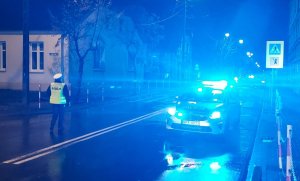 Noc, droga oświetlona. Na jezdni stoi radiowóz z włączonymi światłami błyskowymi w tle policjanci pracujący na miejscu.