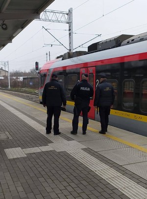 Na zdjęciu na dworcu kolejowym umundurowany Policjant oraz dwóch umundurowanych funkcjonariuszy Straży Ochrony Kolei. W tle pociąg pasażerski.