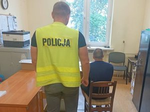 Policjant w kamizelce stoi tyłem przed nim zatrzymany siedzi na krześle.