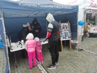 stoisko policyjne na pikniku. Namiot koloru niebieskiego, który zajmuje całość obrazu. W tle widoczny policjant w mundurze z policjantką podczas rozmowy z dwójka dzieci i dorosłym.