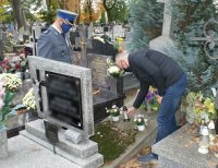 Policjant w mundurze obok niego mężczyzna. Obaj stoją przed grobem. Policjant patrzy na pomnik a mężczyzn składa zapalony znicz na pomnik. W tle inne nagrobki.