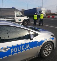 na pierwszym planie bok radiowozu z napisem policja dalej samochód typu bus Inspekcji Transportu Drogowego tuż obok niego dwóch funkcjonariuszy umundurowanych. W tle widoczny samochód ciężarowy.