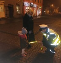 policjant w mundurze wręcza dziecku odblask. Obok opiekunka dziecka. W tle budynki - pora wieczorowa.