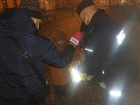 Policjant w mundurze wręcza dziecku odblask. Obok opiekunka dziecka. Reporter radiowy z mikrofonem. W tle budynki - pora wieczorowa.