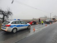 Radiowóz- przed nim policjanci pracujący na miejscu wypadku. Dwa pojazdy rozbite. Jeden na jezdni drugi poza nią. W tle budynki i pola oraz inne auto.