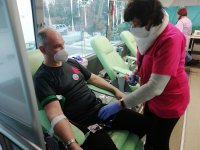 Na zdjęciu pielęgniarka podłącza aparaturę do poboru krwi siedzącemu na fotelu mężczyźnie ubranemu w czarną koszulkę z czerwoną krwinką. Mężczyzna ma zasłonięty nos i usta maseczką ochronną koloru białego.