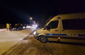 Policjanci podczas kontroli osobowego auta. W tle jadące pojazdy i stojący na poboczu radiowóz.