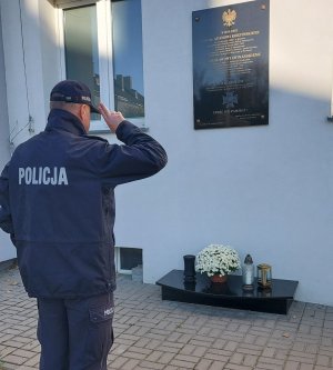 Policjant oddaje honor przed tablicą pamiątkową wmurowaną w ŚCIANĘ KOMENDY POLICJI.