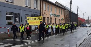 Policjanci, uczestnicy wydarzenia ubrani w kamizelki odblaskowe z banerami dotyczącymi promocji bezpieczeństwa na drodze idą ulicą.