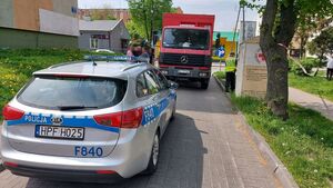 Na drodze, Przed radiowozem stoi auto ciężarowe. Obok umundurowany policjant. W tle bloki mieszkalne, drzewa i inne zabudowania.