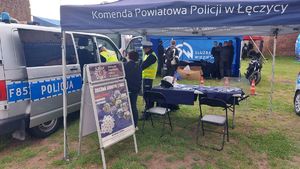 Stoisko promocyjne policji na pikniku motocyklowym. Policjant stoi obok radiowozu z uczestnikami wydarzenia.