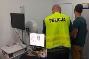 Policjant w kamizelce z napisem POLICJA z zatrzymanym podczas daktyloskopii stoją tyłem.