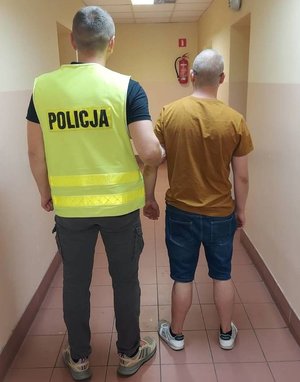 Zdjęcie przedstawia policjanta w odblaskowej kamizelce z napisem POLICJA wraz ze stojącym obok niego mężczyzną w pomarańczowej koszulce i jeansowych spodniach.