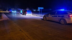 Zdjęcie przedstawia miejsce wypadku drogowego po zmroku. Na jezdni usytuowany jest samochód osobowy biorący udział w zdarzeniu oraz policyjny radiowóz oznakowany. Obok umundurowany funkcjonariusz oraz mężczyzna w kamizelce odblaskowej trzymający walizkę.