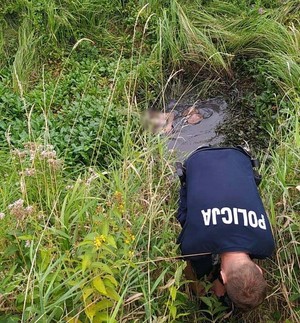 Rów porośnięty trawą i wypełniony woda, obok policjant wchodzi do rowu.