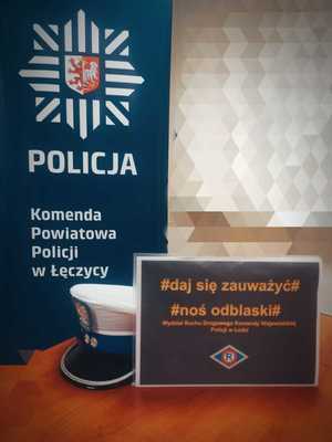 Zdjęcie przedstawia plakat akcji #daj się zauważyć#, #noś odblaski# na tle baneru Komendy Powiatowej Policji w Łęczycy i policyjnej czapki z białym otokiem.