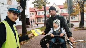 Zdjęcie przedstawia mężczyznę z dzieckiem, któremu umundurowany policjant wręcza kamizelkę odblaskową.