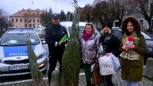 Policjant z redaktorką radiaq rozmawiają z mieszkańcami i wręczają choinki i odblaski, W tle radiowóz i policjanci ruchu drogowego.