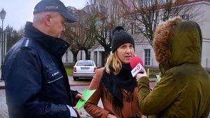 Policjant z redaktorką radiaq rozmawiają z mieszkańcami i wręczają choinki i odblaski, W tle radiowóz i policjanci ruchu drogowego.