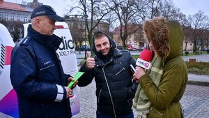 Policjant z redaktorką radiaq rozmawiają z mieszkańcami i wręczają choinki i odblaski, W tle radiowóz i policjanci ruchu drogowego.