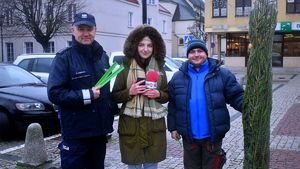 Policjant z redaktorką radiaq rozmawiają z mieszkańcami i wręczają choinki i odblaski, W tle radiowóz i policjanci ruchu drogowego.