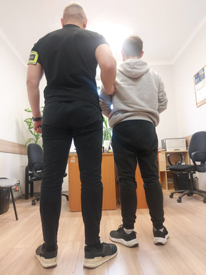 Nieumundurowany policjant z opaską na lewym ramieniu z napisem Policja wraz z zatrzymanym 22-latkiem.
