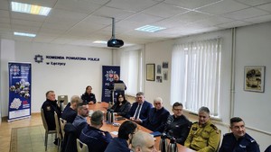 Radiowozy zaparkowane przed komendą. Policjanci w mundurach podczas odprawy.