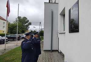 Umundurowani policjanci przed tablicą i pomnikami oddają honory.