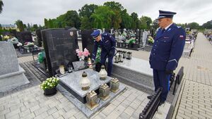 Umundurowani policjanci przed tablicą i pomnikami oddają honory.